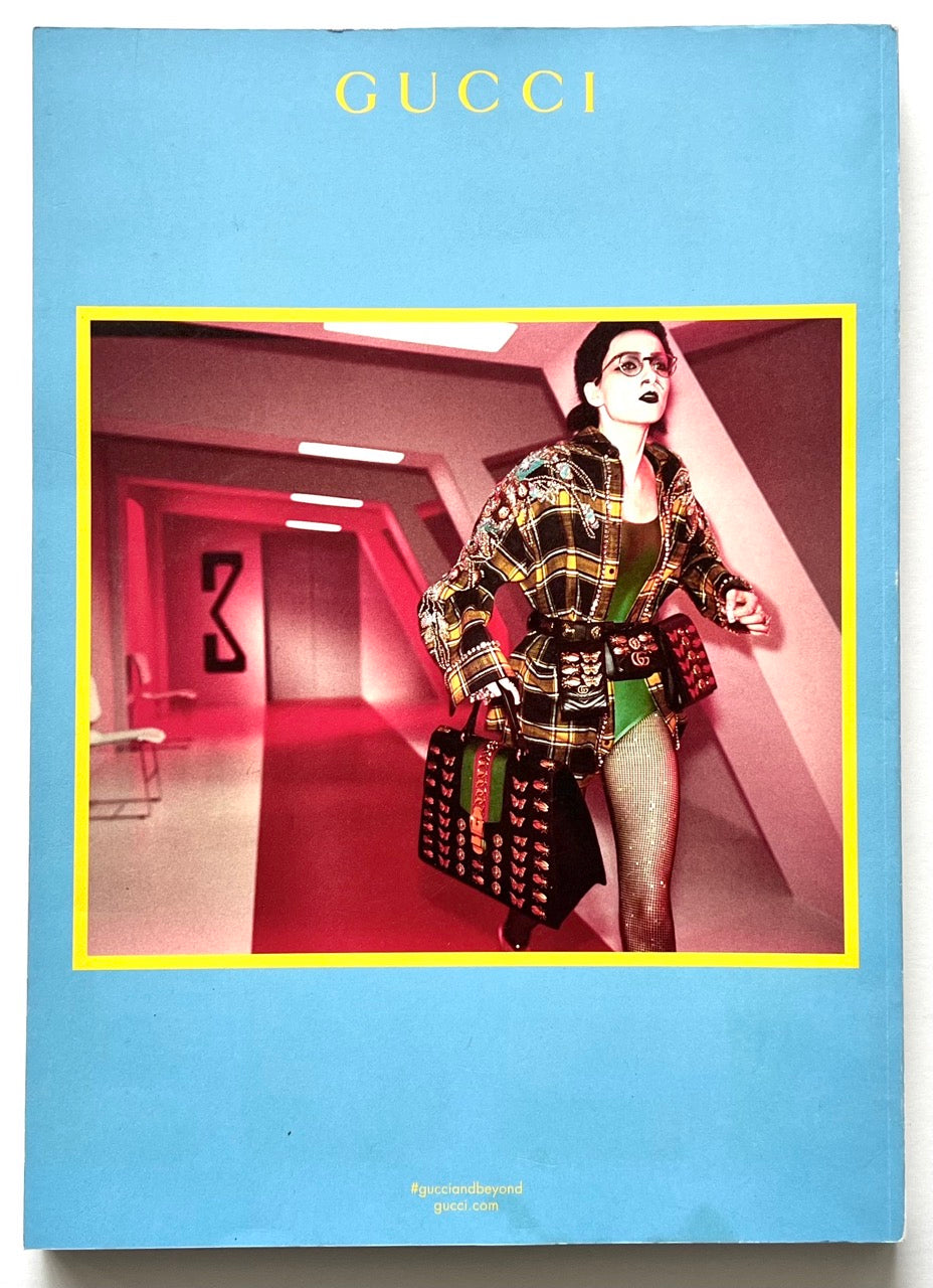 A colourful Gucci advertisement showing a model running through a futuristic corridor lit in red, wearing a yellow plaid jacket, embellished accessories, and carrying a large black Gucci bag, set against a bright blue background with a yellow border.