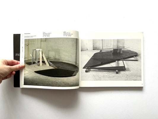Open spread from “The Sculpture Show” (1983) featuring two sculptural installations by Michael Kenny: a colour photograph of geometric wood and metal forms arranged in a studio space on the left, and a black-and-white photograph of a large angular floor sculpture on the right.