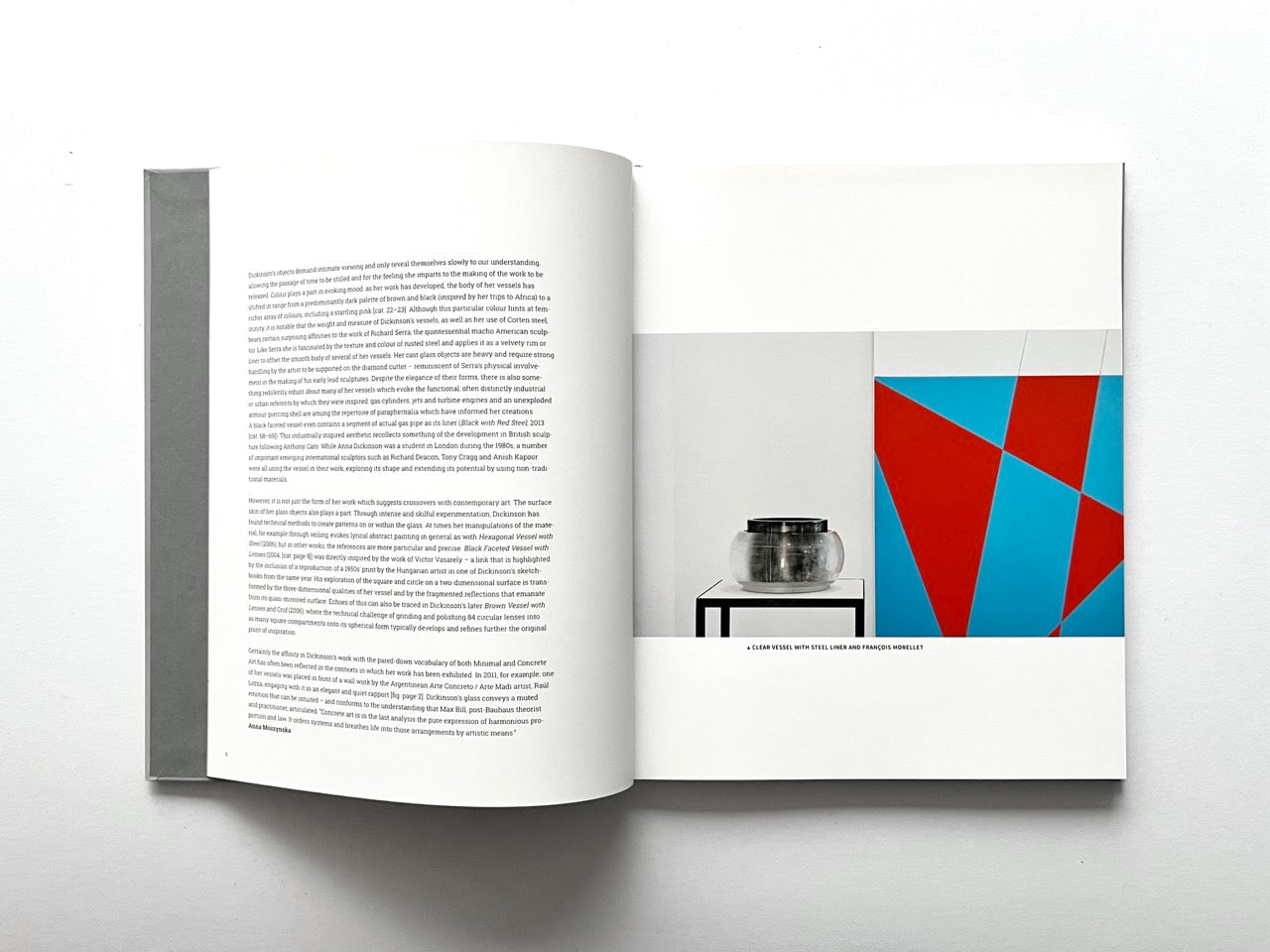 Open spread of the book “Anna Dickinson”, showing a page of essay text on the left and a full-page image on the right combining a clear glass vessel with a geometric red and blue abstract background, held open against a white background.