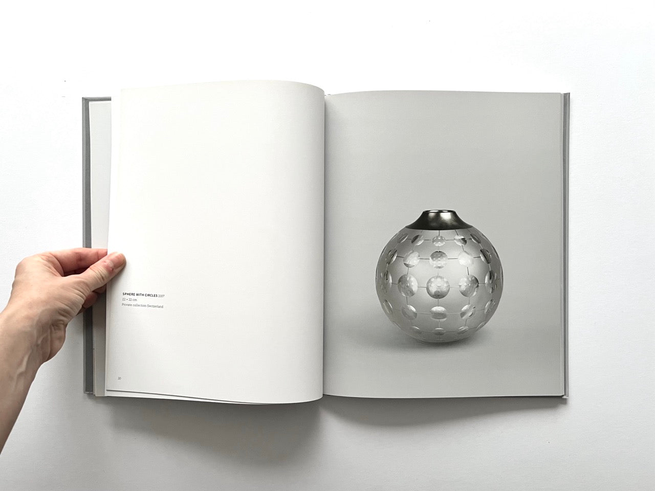 Open spread of the book “Anna Dickinson”, showing a full-page photograph of a spherical clear glass vessel with circular cut-out details and a dark rim on the right page, against a neutral grey background.