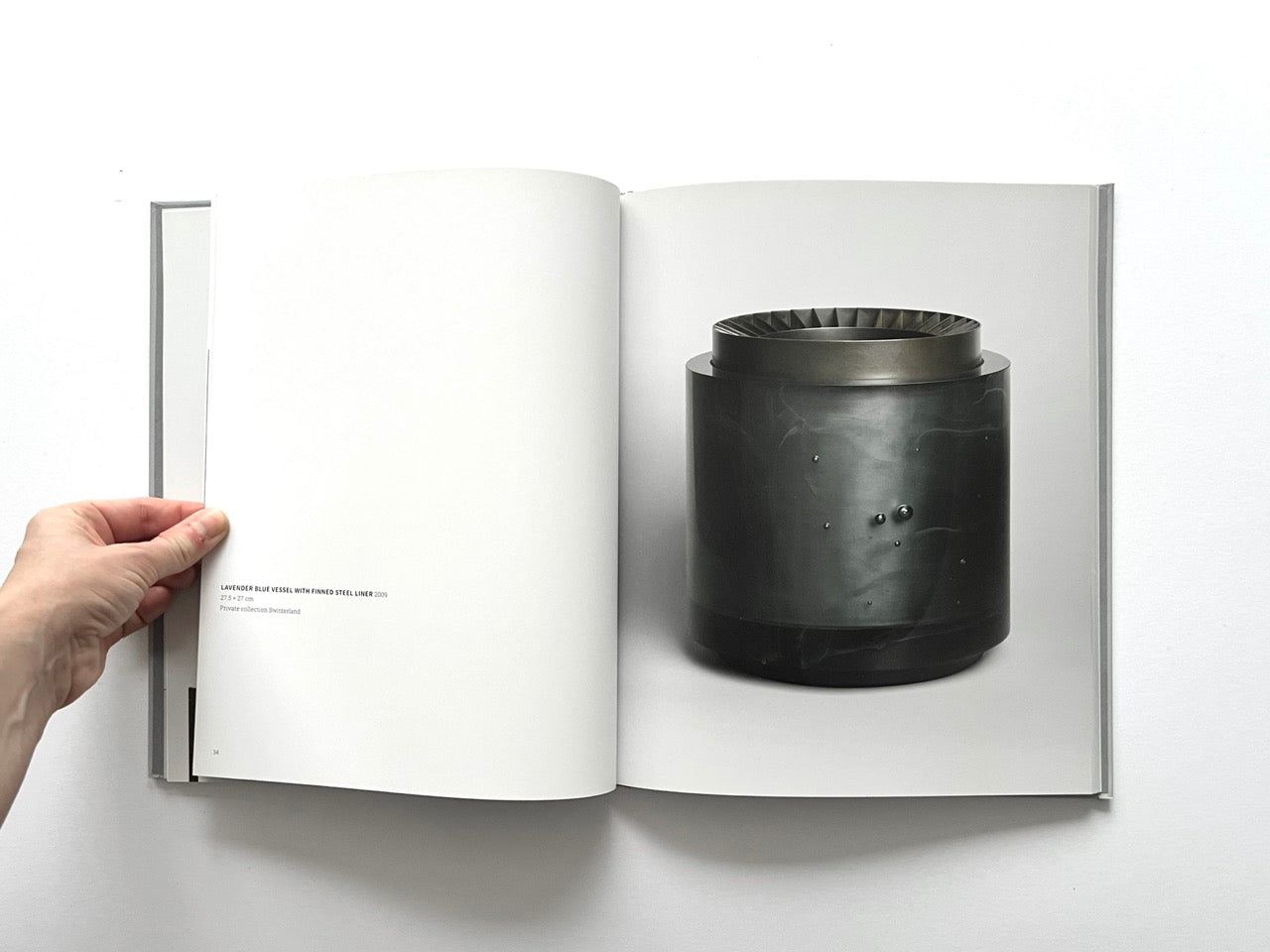 Open spread of the book “Anna Dickinson”, showing a full-page photograph of a dark cylindrical glass vessel with a steel-lined rim and subtle surface details on the right page, against a neutral grey background.