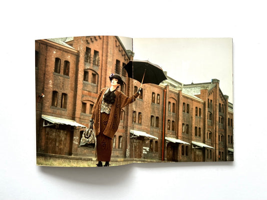 Interior spread from Atsuki Onishi's Knit Picture Book, published in 1989, featuring a fashion model in an elaborate brown knit ensemble posing with an umbrella outside historic red brick warehouse buildings.