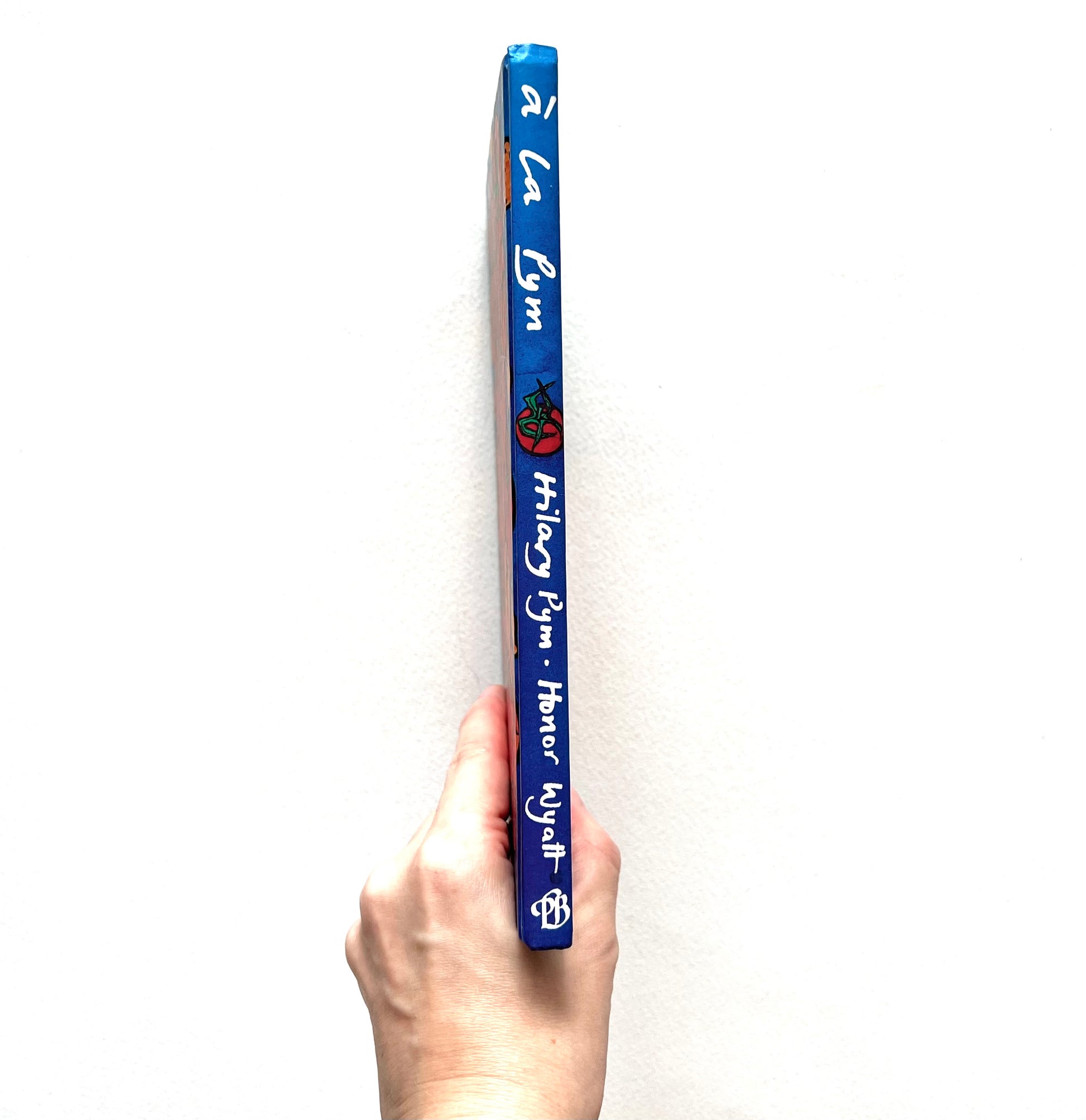 Spine-view of 'A La Pym' cookery book held by a hand on a white background