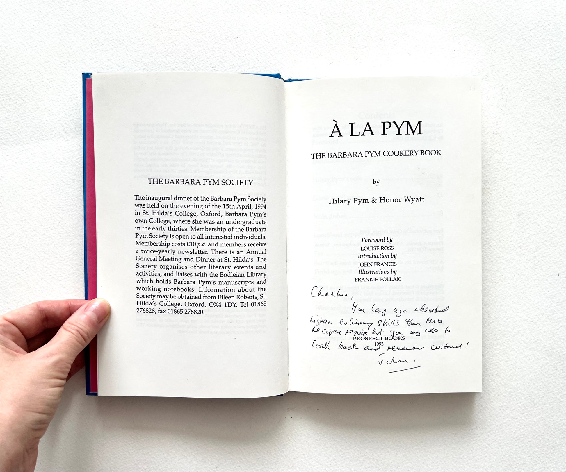 Open book showing a title page of 'À La Pym' cookery book held by a hand on a white background