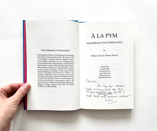 Open book showing a title page of 'À La Pym' cookery book held by a hand on a white background