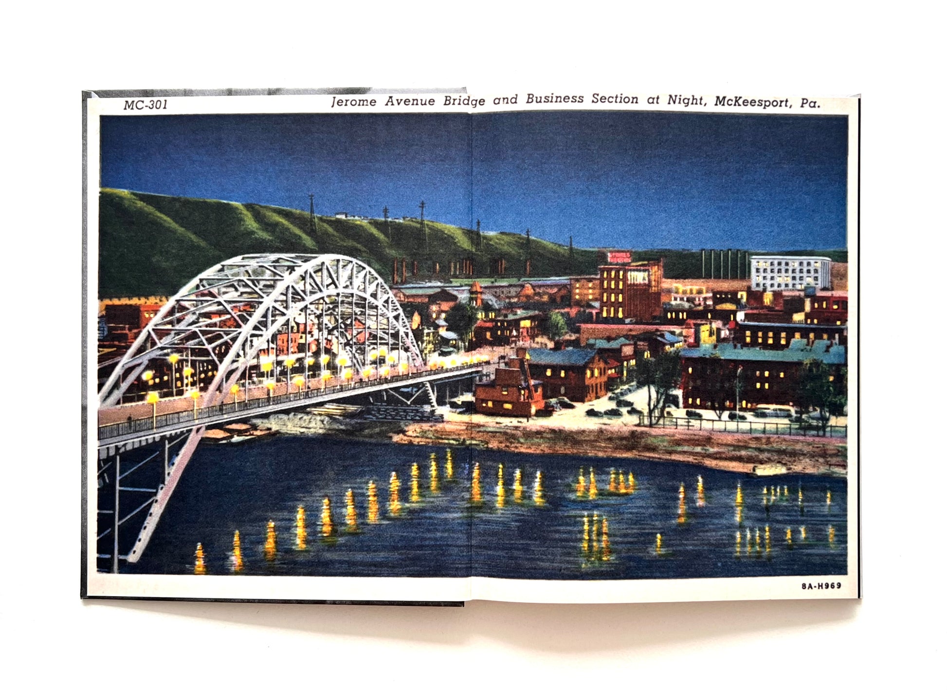 Open book showing endpapers featuring an enlarged vintage-style postcard of a bridge and cityscape of McKeesport at night, featuring illuminated buildings and a river.