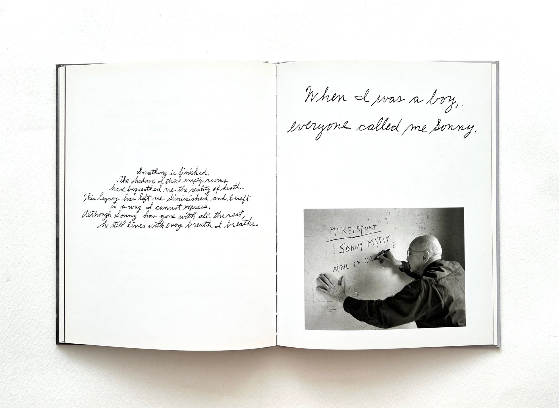 Open book with handwritten text and a black and white photo of a bold man writting on a wall on a white background