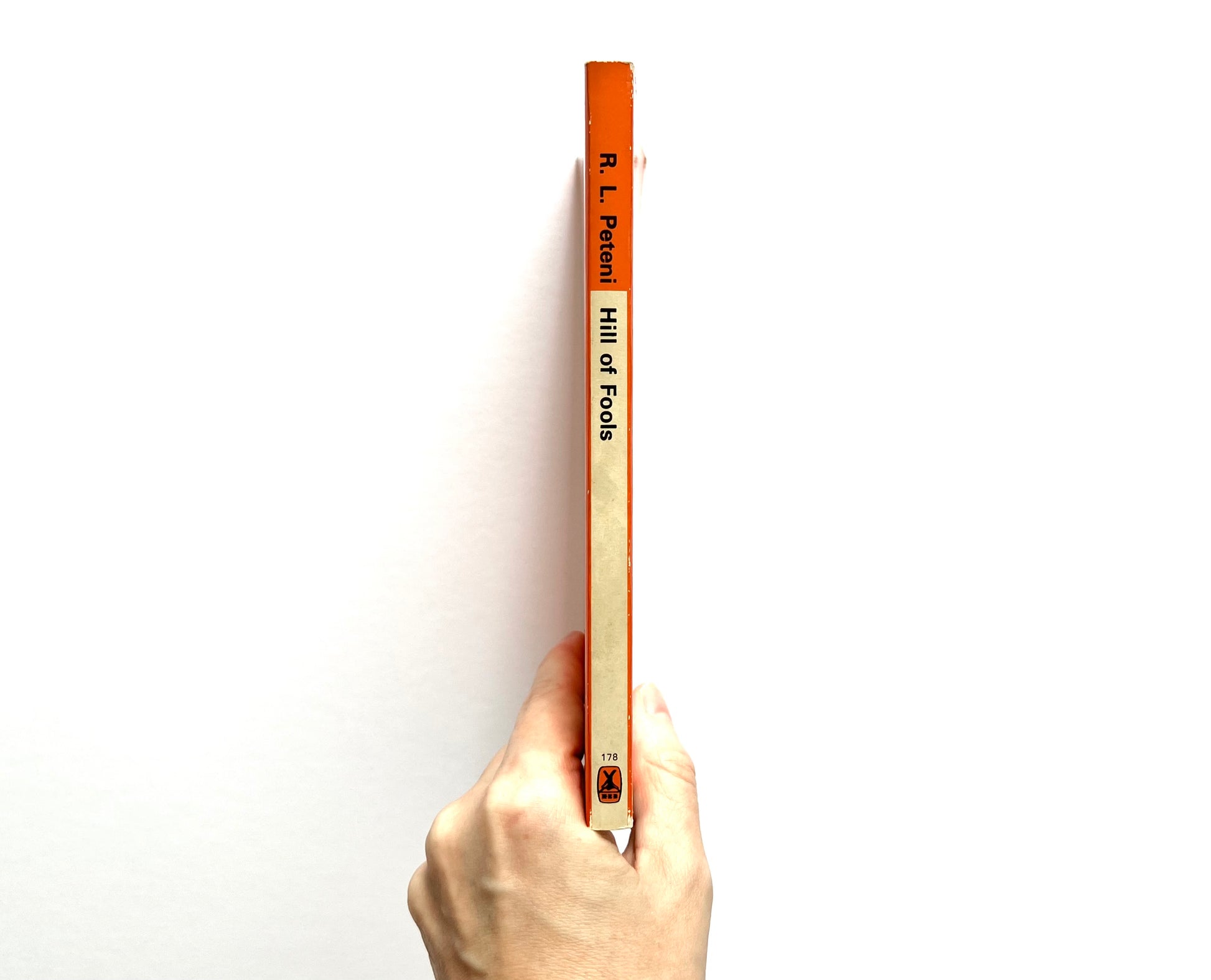 Hand holding a book titled 'Hill of Fools' by R.L. Peteni against a white background