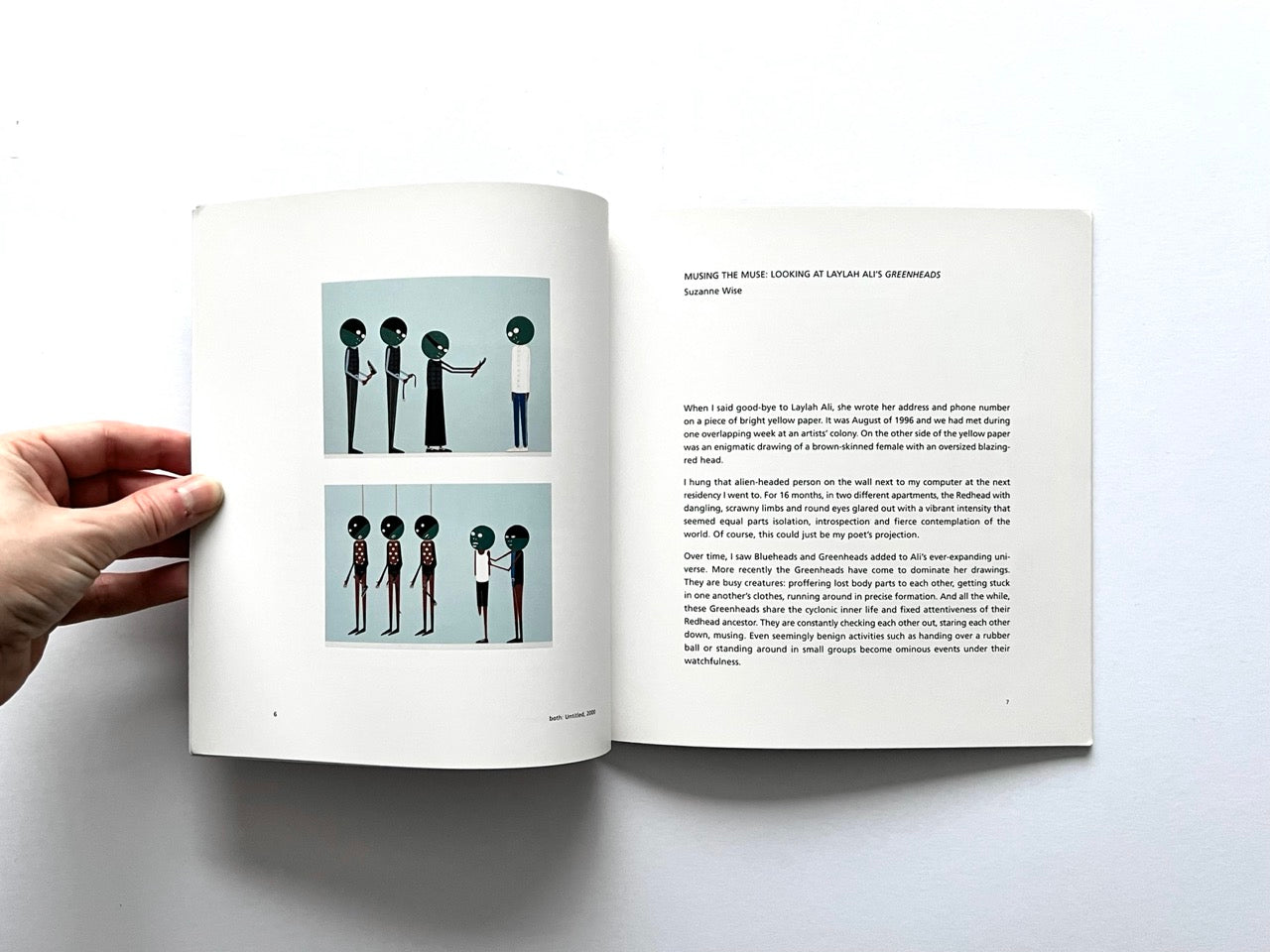 Open spread of the book “Laylah Ali”, showing two small reproductions of figurative paintings with stylised human figures on the left page and an essay titled “Musing the Muse: Looking at Laylah Ali’s Greenheads” on the right page, held open by a hand against a white background.