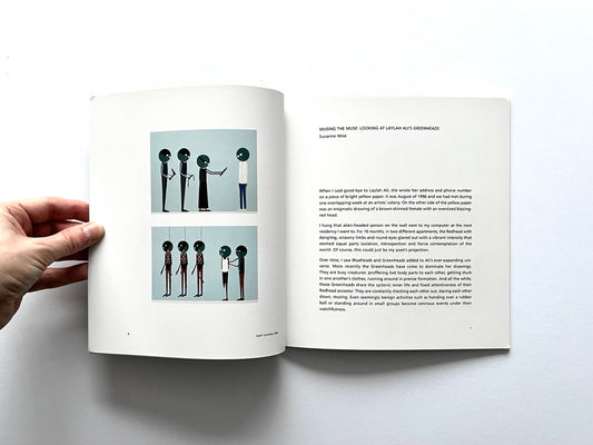 Open spread of the book “Laylah Ali”, showing two small reproductions of figurative paintings with stylised human figures on the left page and an essay titled “Musing the Muse: Looking at Laylah Ali’s Greenheads” on the right page, held open by a hand against a white background.