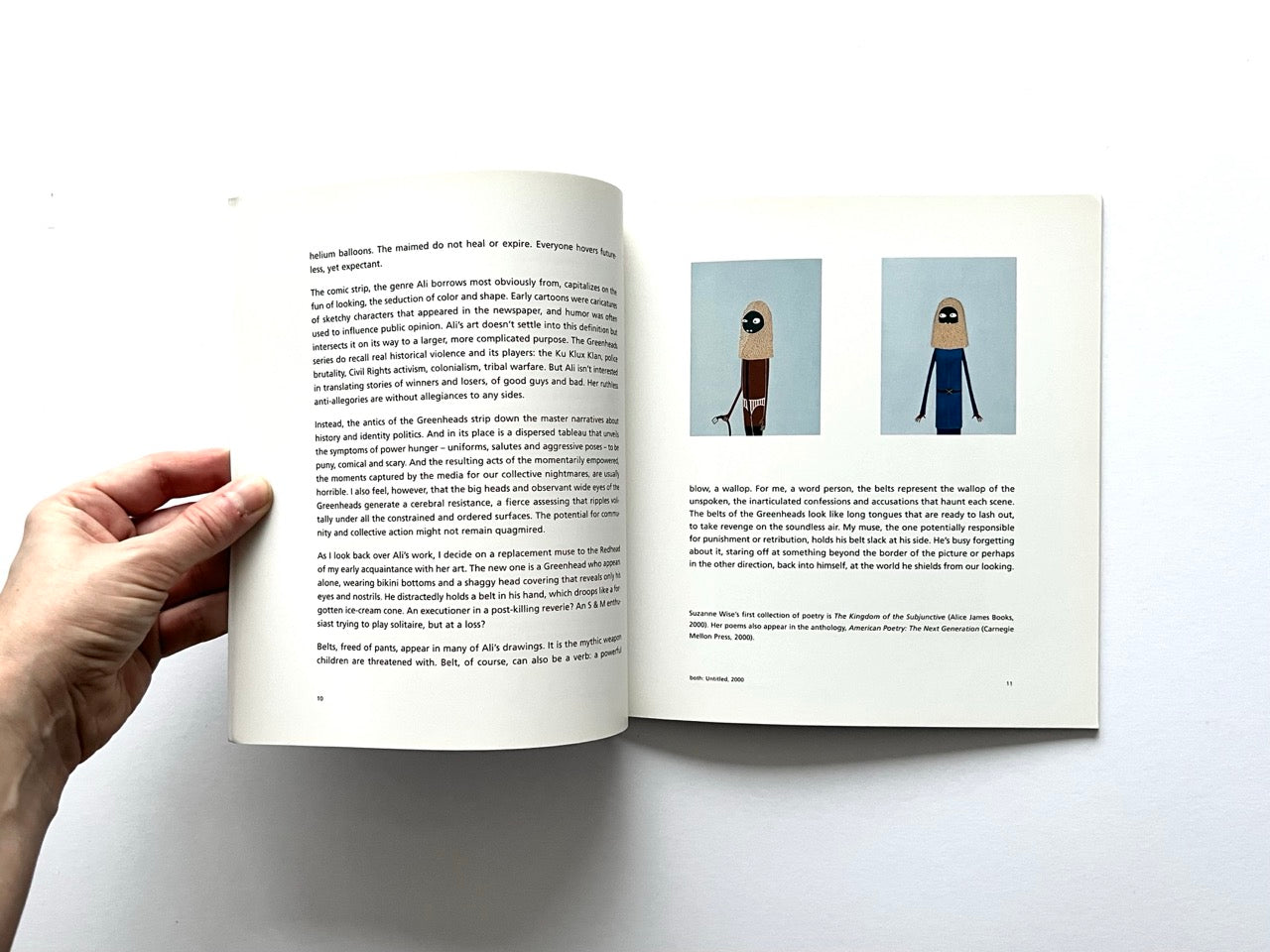 Open spread of the book “Laylah Ali”, showing a page of essay text on the left and two small reproductions of figurative paintings with stylised human figures on pale blue backgrounds on the right, held open by a hand against a white background.