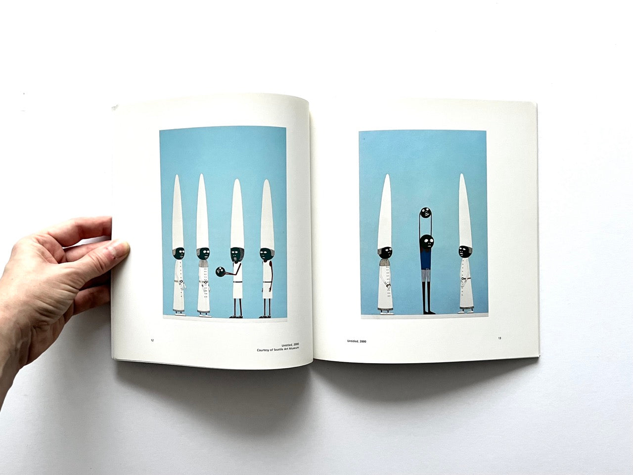Open spread of the book “Laylah Ali”, showing two full-page reproductions of figurative paintings with stylised human figures wearing tall white pointed hats against pale blue backgrounds, held open by a hand against a white background.