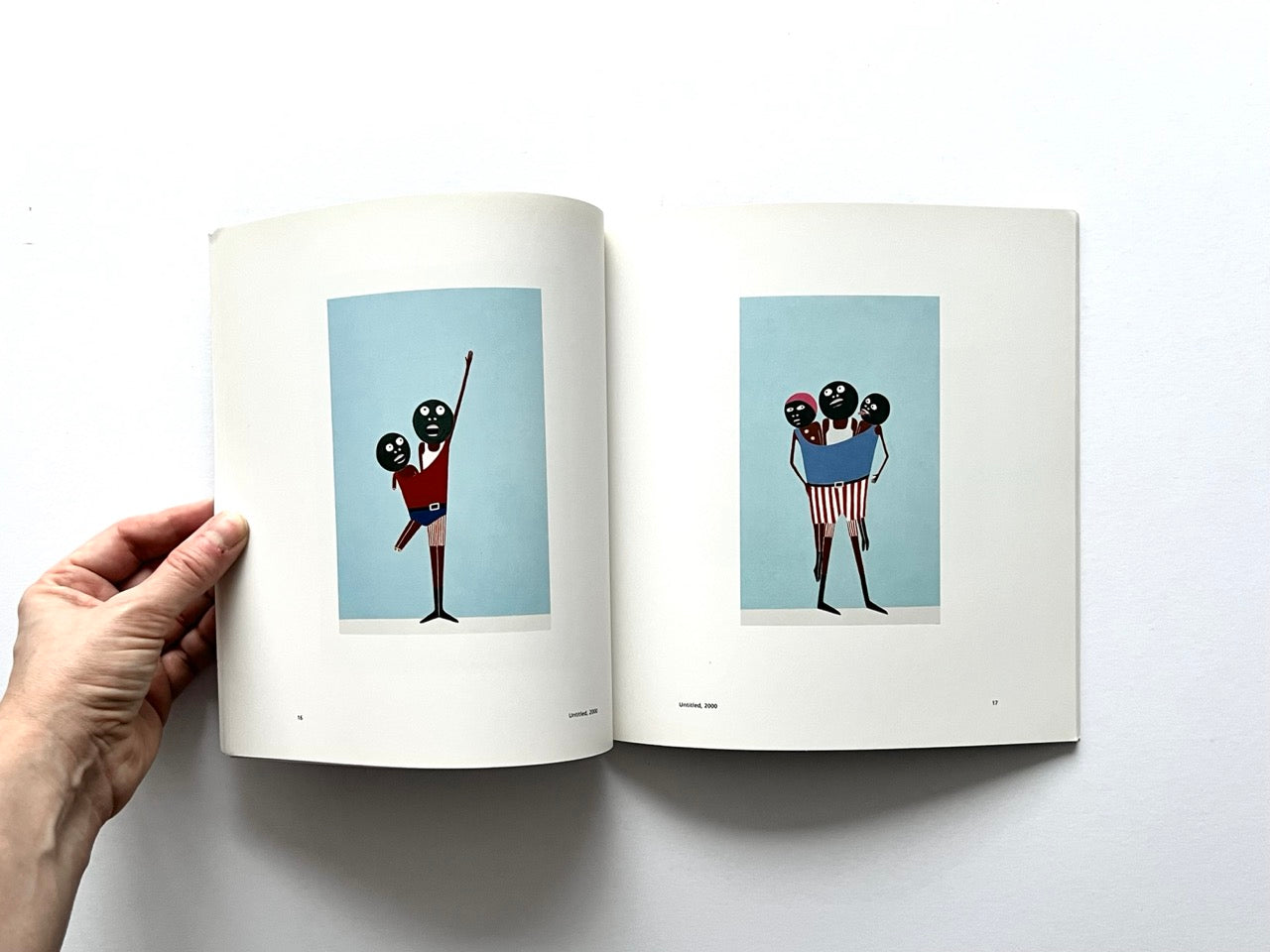 Open spread of the book “Laylah Ali”, showing two full-page reproductions of figurative paintings with stylised human figures on pale blue backgrounds, one depicting a raised arm gesture and the other a grouped figure composition, held open by a hand against a white background.