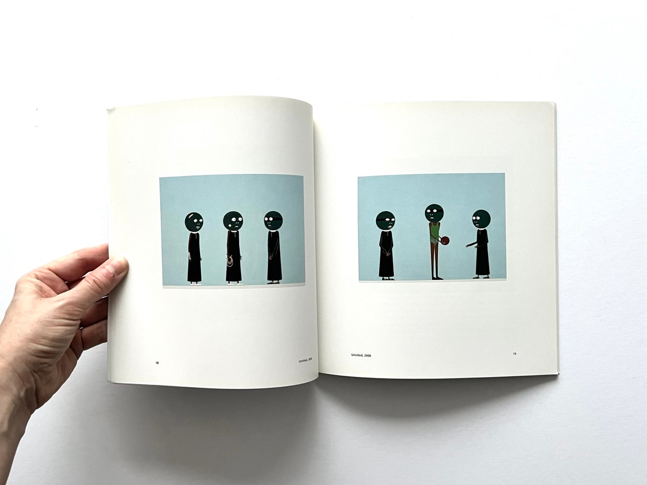 Open spread of the book “Laylah Ali”, showing two small reproductions of figurative paintings with stylised human figures in dark clothing against pale blue backgrounds, held open by a hand against a white background.