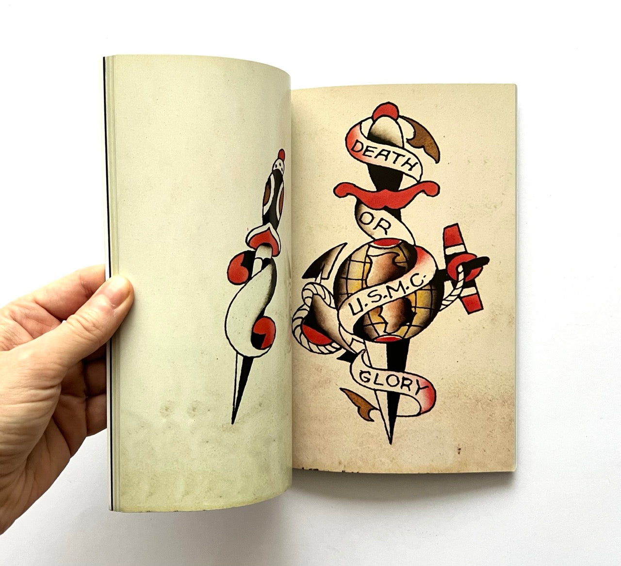 Open book displaying a traditional tattoo design with an anchor, crossed swords, and text on a beige background.