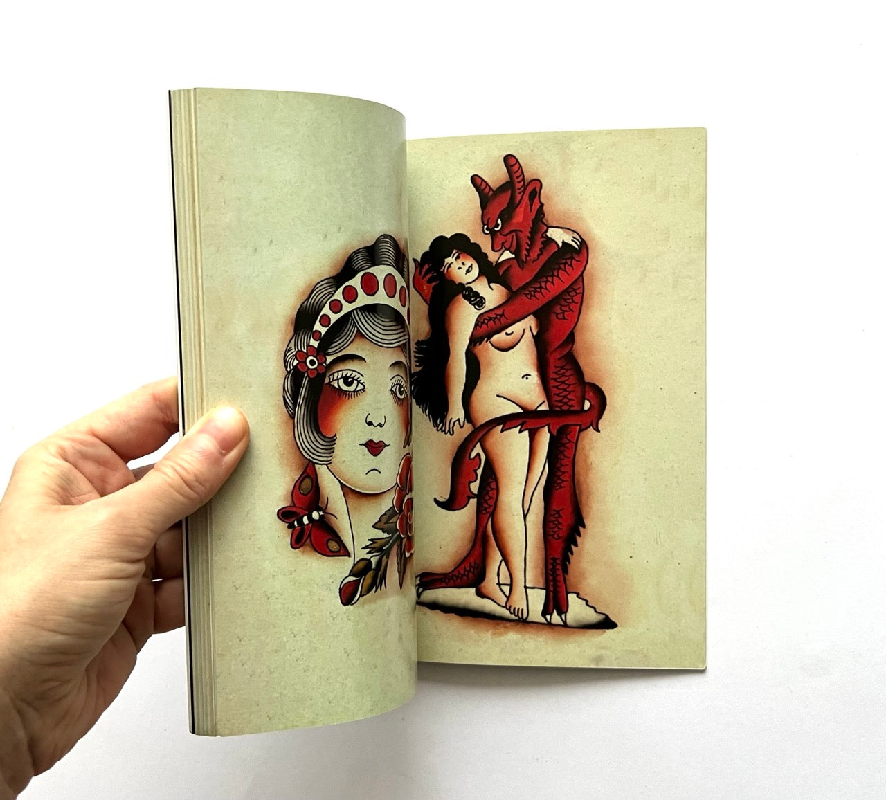 Open book - 'LOST ART: FROM TATTOOING’S PAST' - displaying vintage tattoo designs of a naked woman embraced by the devil on a beige background.