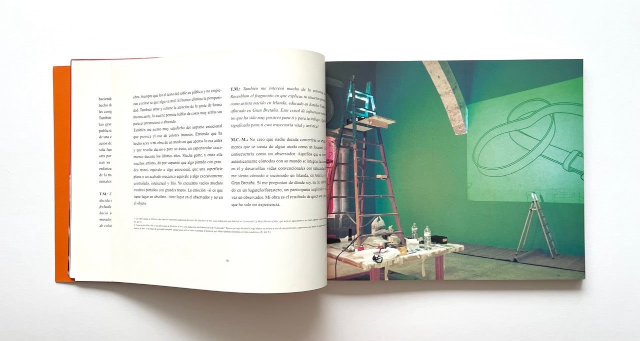 Open book with text on one page and a photograph of a gallery exhibition setup on the other, on a white background.