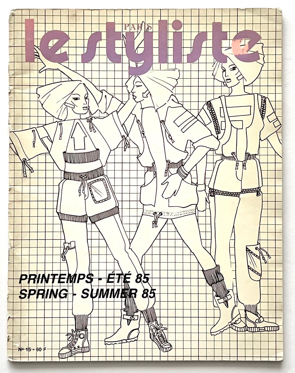 Front cover of Paris Le Styliste No 15 Spring–Summer 1985: cream grid background with three outlined fashion illustrations in sporty 80s silhouettes, oversized hoods and layered garments; bold lavender title at top; French/English season text printed in black at bottom.