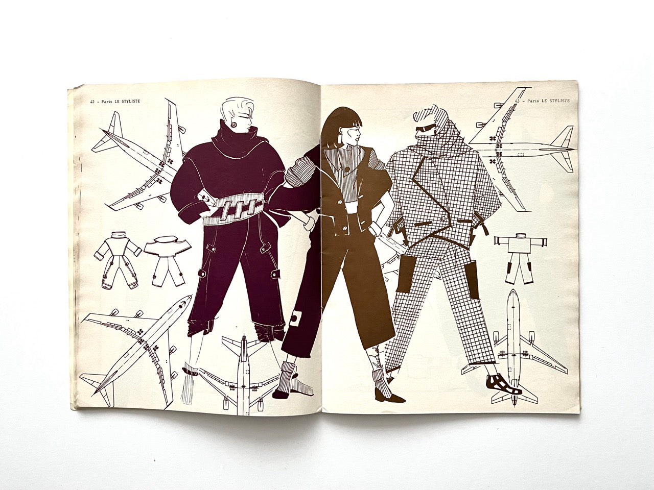 Two-page spread from Paris Le Styliste No. 15 (1985) displaying three figures in aviation-inspired outerwear: oversized padded jackets, cropped trousers and checked coats; repeated airplane line drawings surround the models alongside small technical sketches.