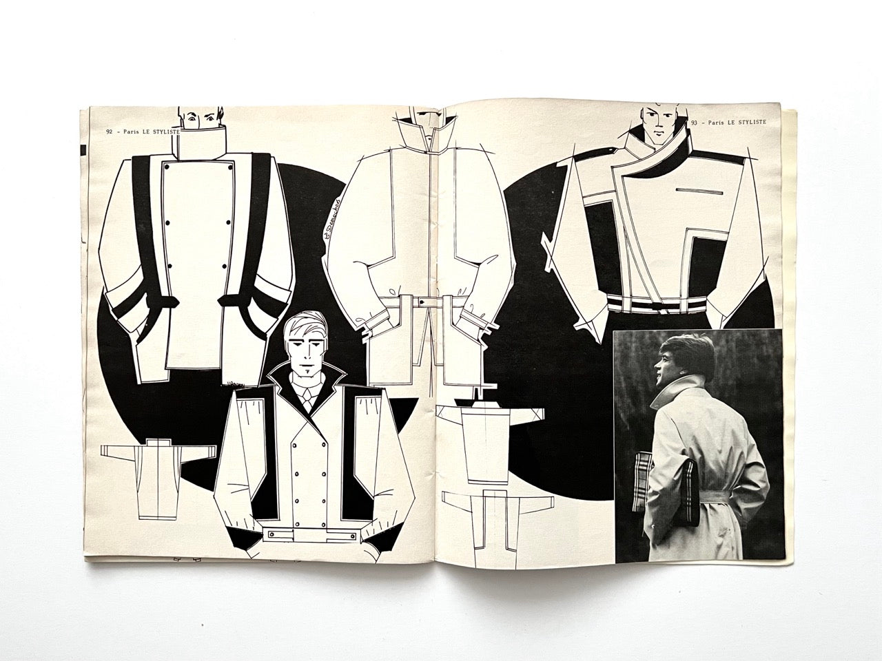 Spread from Paris Le Styliste No. 15 (1985) showing angular menswear coat designs with wide lapels, double-breasted fronts and graphic black paneling; inset black-and-white photograph of a man in a belted trench coat holding a folded newspaper.