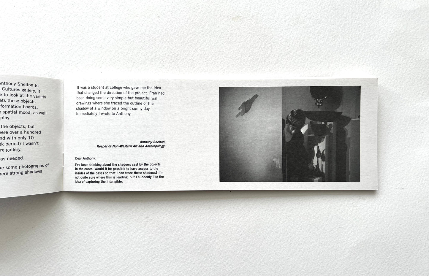 Open book with text and a black and white photograph on a white wall.