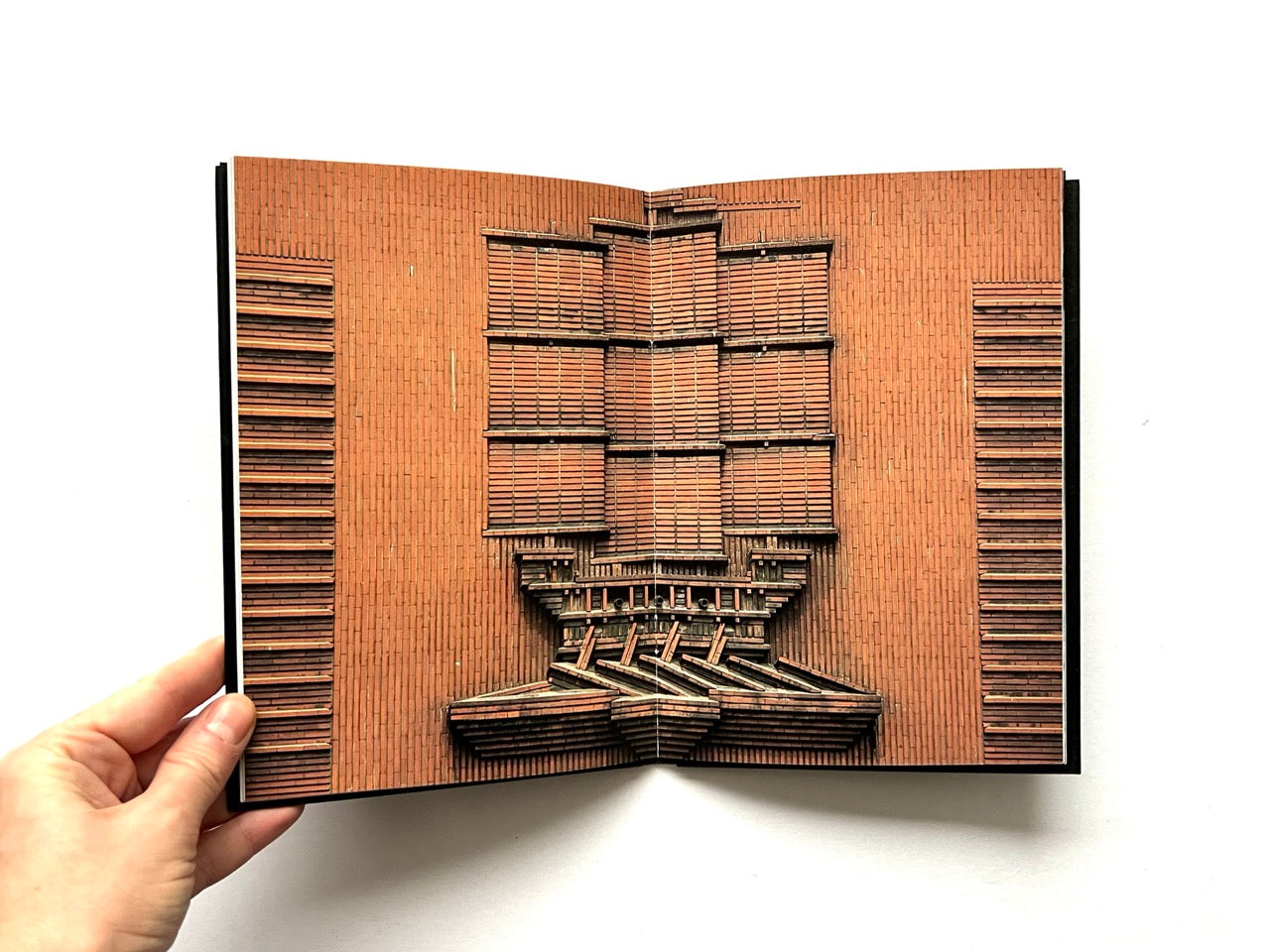 Open spread of Pentagram Papers 24 “The Arms of Paris”, showing a full-bleed photograph of a symmetrical brick architectural facade with layered horizontal elements, held open by a hand against a white background.