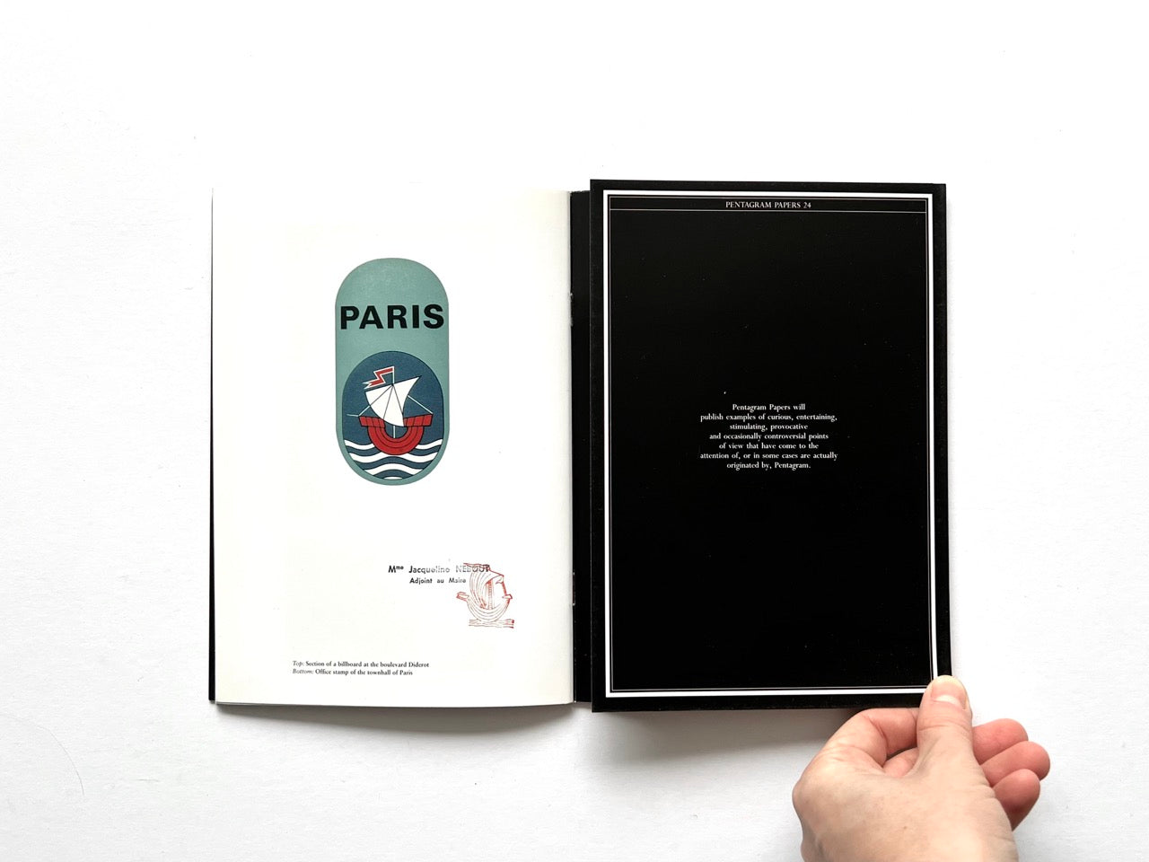 Open spread of Pentagram Papers 24 “The Arms of Paris”, showing a white page with a Paris emblem featuring a stylised ship on the left and a black colophon page with centered text and fine white border on the right, held open by a hand against a white background.