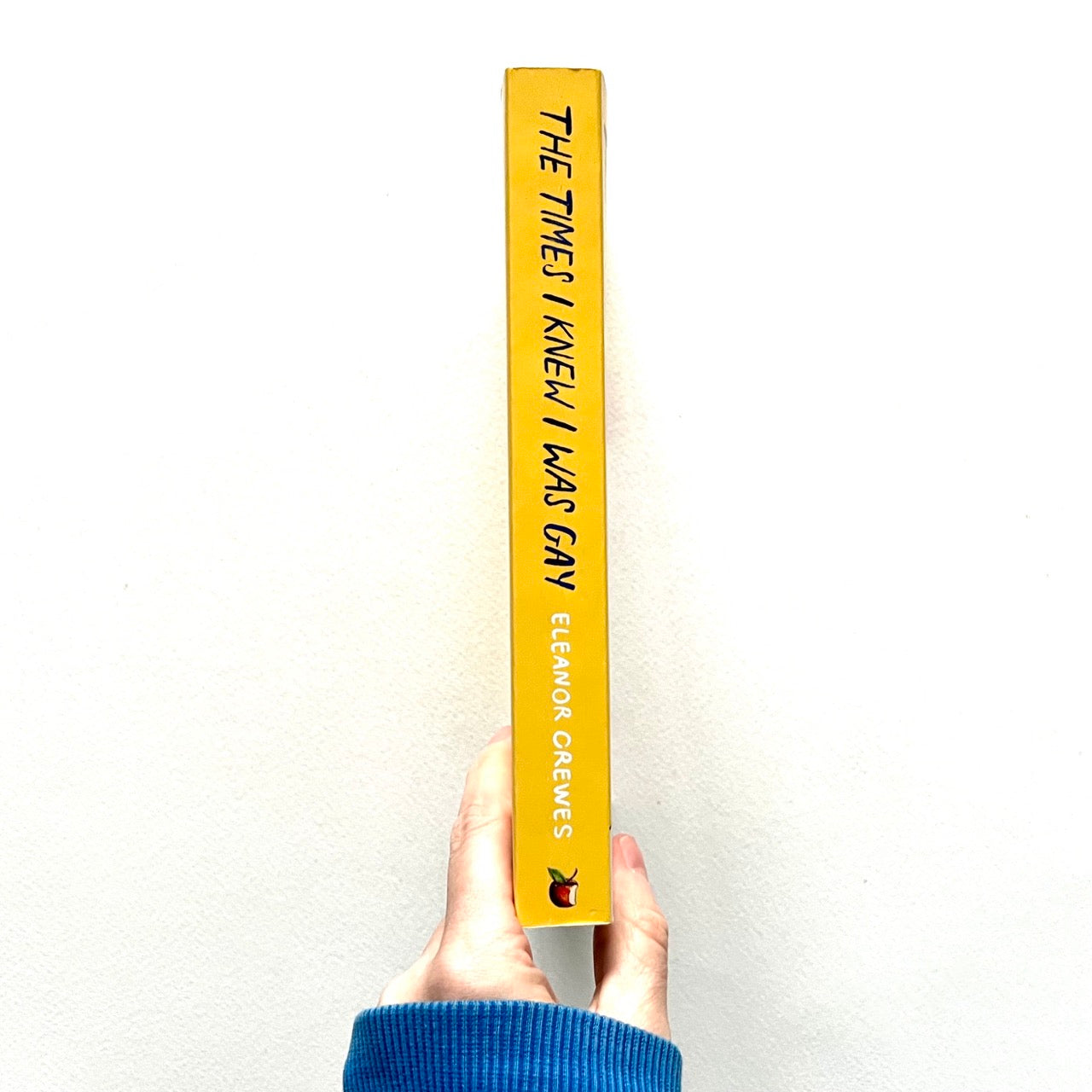 Spine-view of book titled 'The Times I Knew I Was Gay' by Eleanor Greves, held by a hand with a blue sleeve on a white background