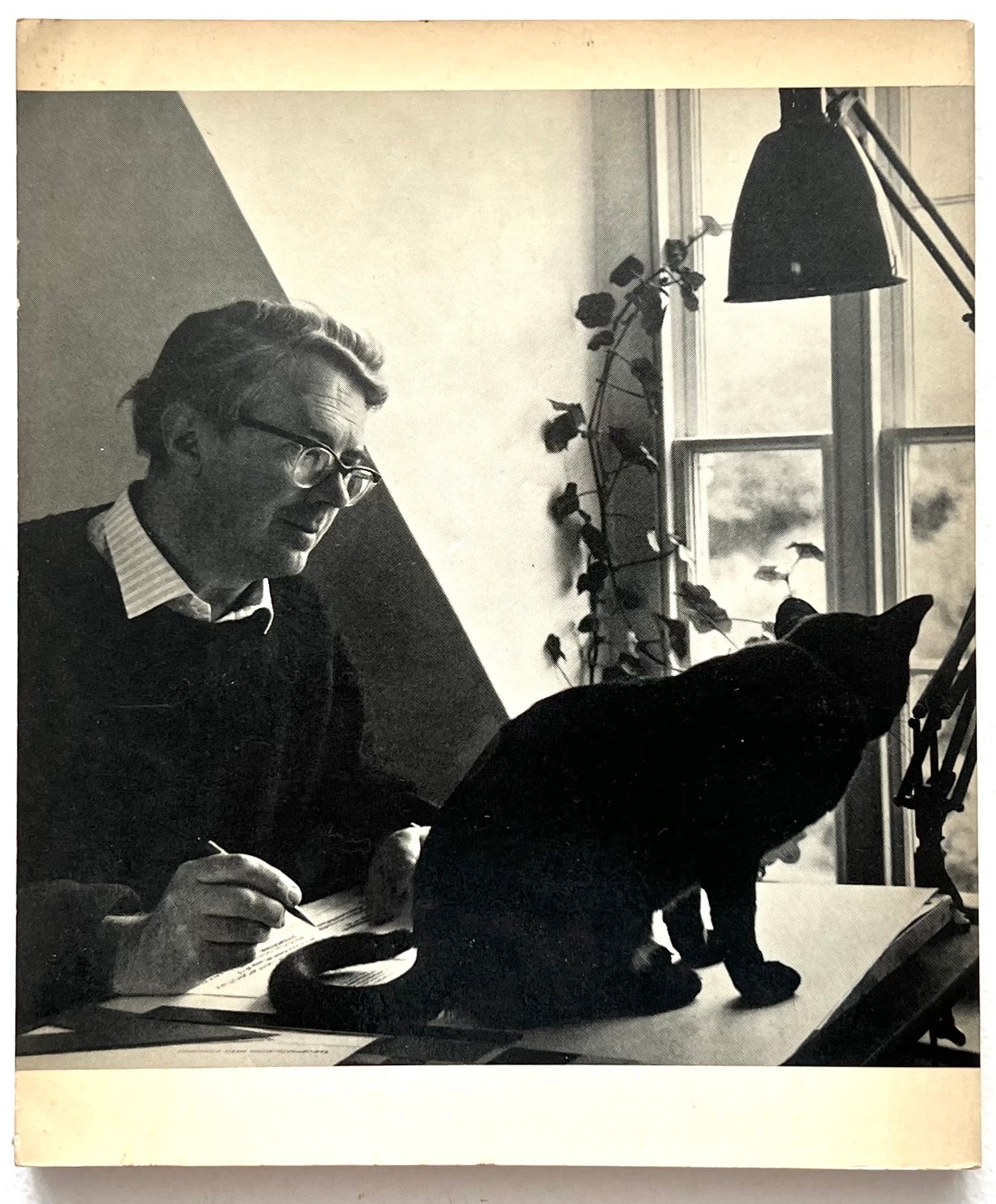 Rear cover of 'Typography: basic principles' by John Lewis showing a photo of the author writing at a desk with a black cat sitting on top of it, in a room with a lamp and window.