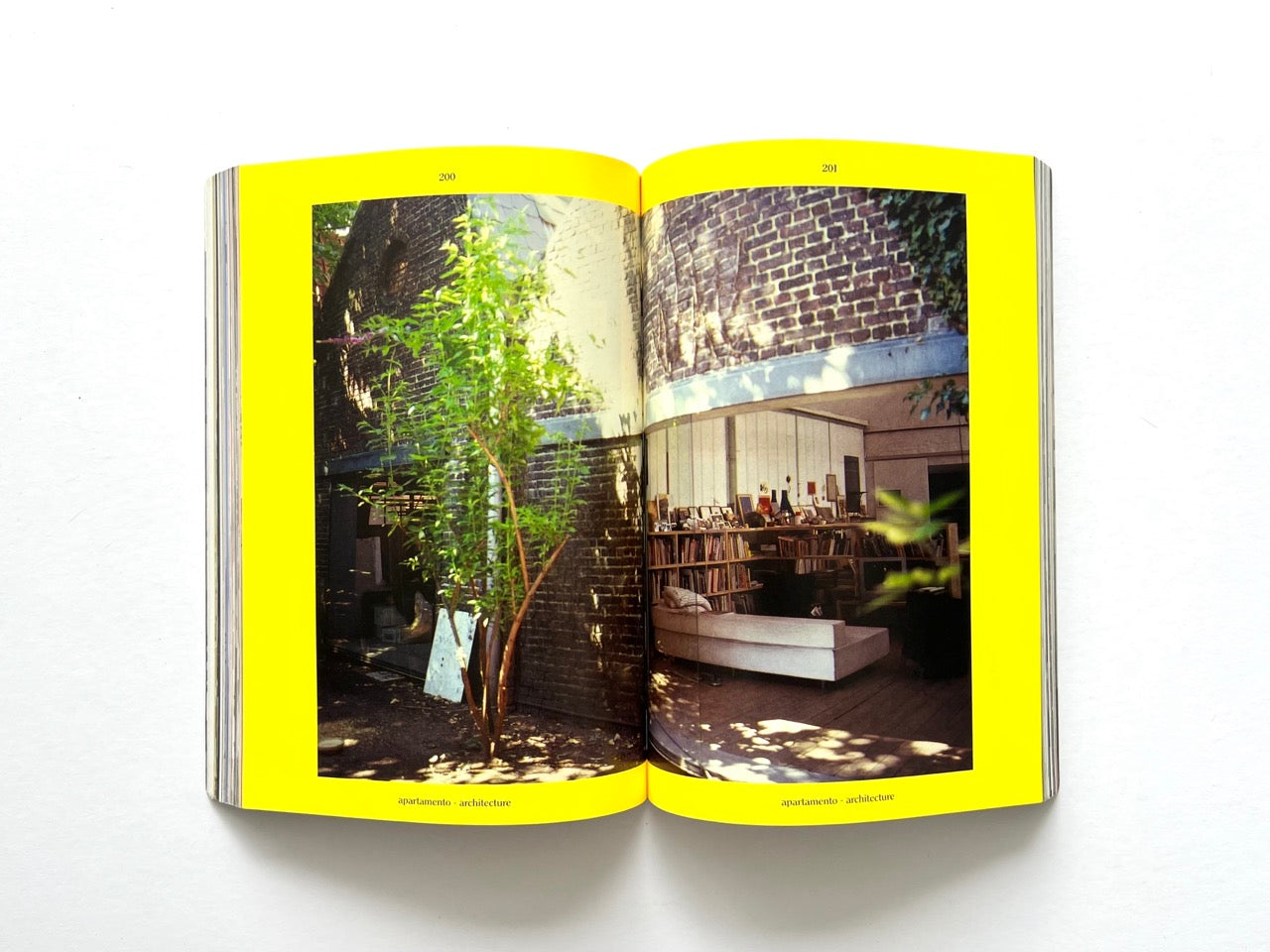 Apartamento Magazine Issue #21 – Interior spread showing a brick courtyard and adjacent living space with bookshelves, sofa, and tree, framed by bright yellow pages.