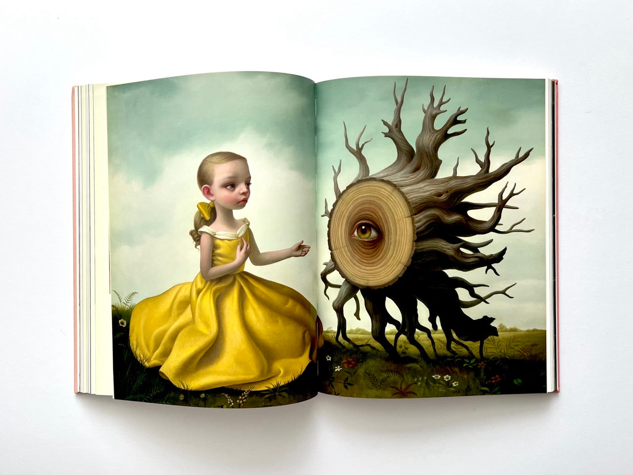 Mark Ryden Pinxit interior spread showing a girl in a yellow dress facing a fantastical tree trunk with a single eye at its center in a soft landscape setting