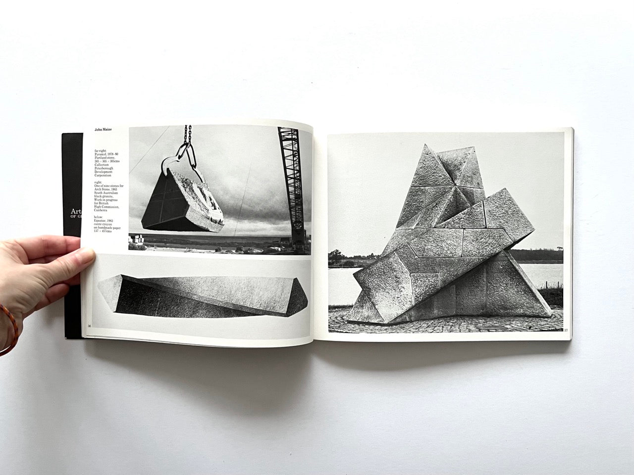 Open spread from “The Sculpture Show” (1983) showing black-and-white photographs of large geometric stone sculptures by John Maine, including a block being lifted by crane and a multi-part outdoor installation beside water.