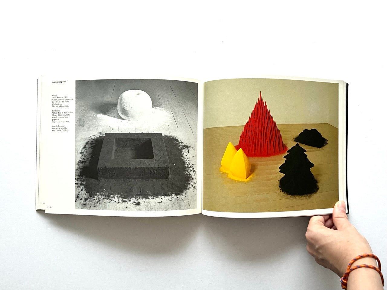Open spread from “The Sculpture Show” (1983) showing a black-and-white photograph of a square earth pigment sculpture and rounded form on the left, and a colour photograph on the right of bright red, yellow and black pigment-based sculptural mounds - both by Anish Kapoor.